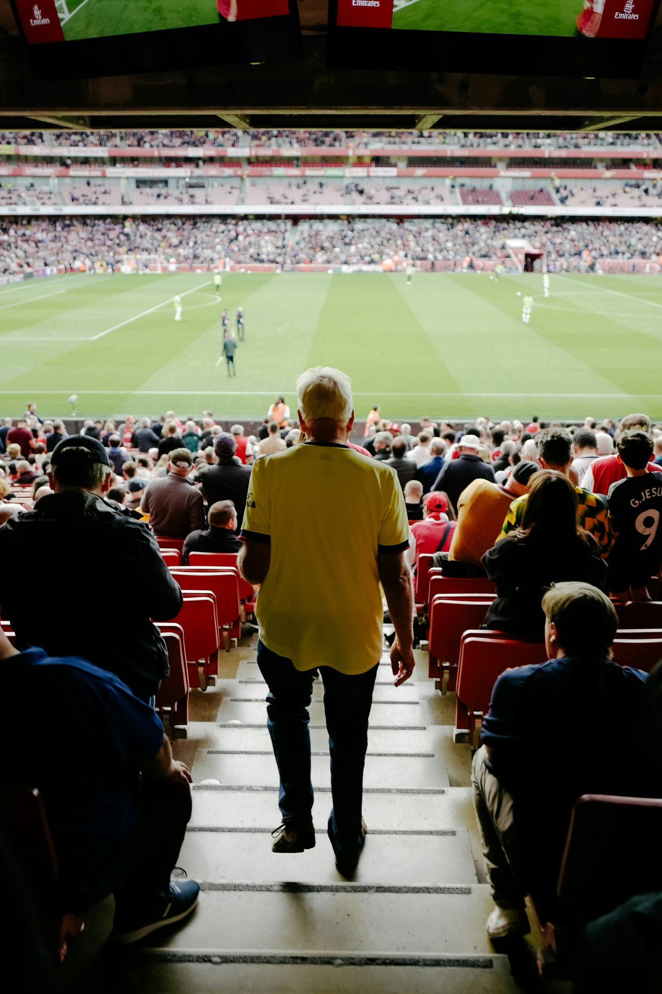 An older supporter in a yellow shirt walking down the steps of a stand towards the pitch