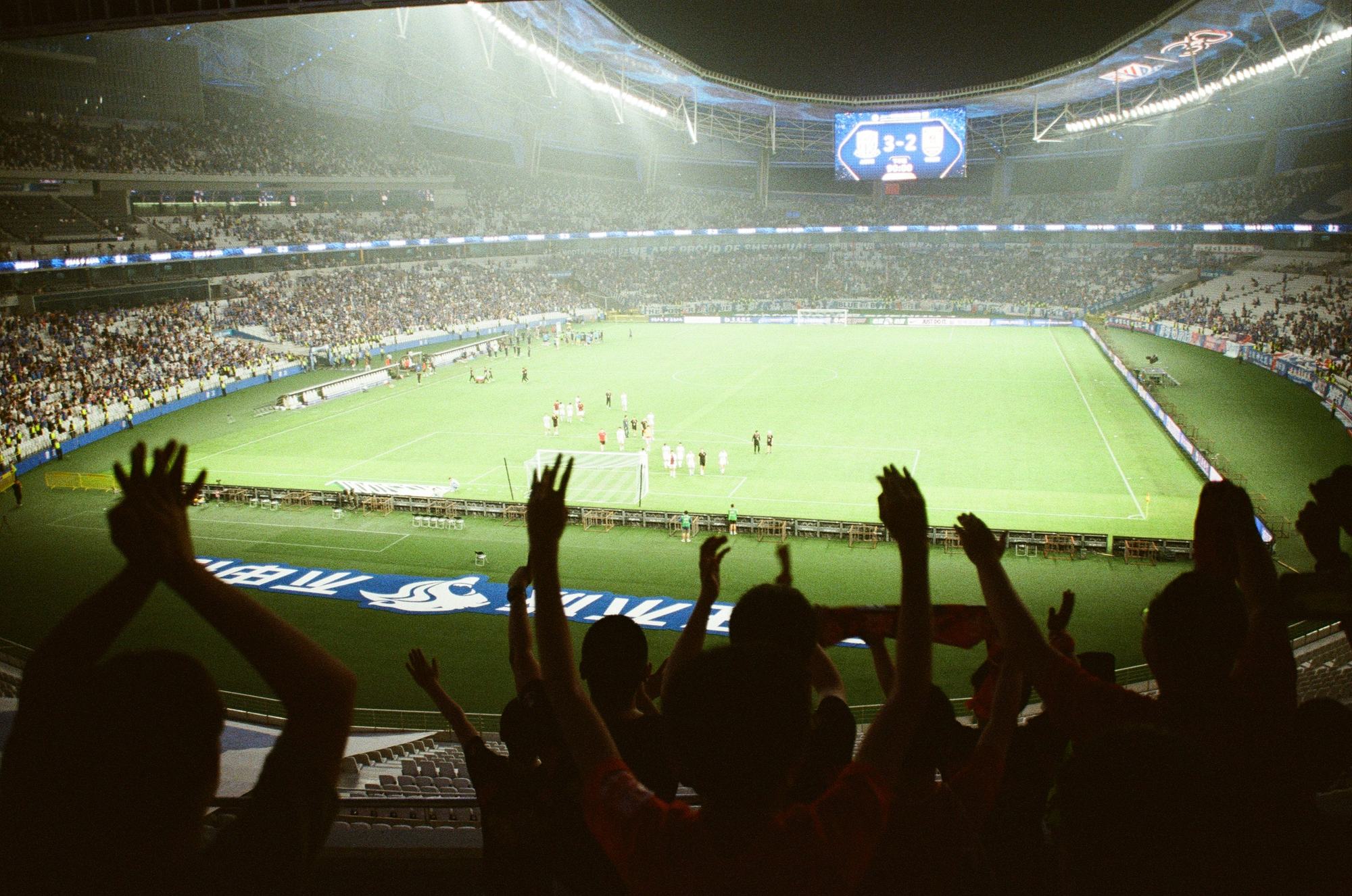 Supporters with their arms raised, silhouetted against the floodlit pitch